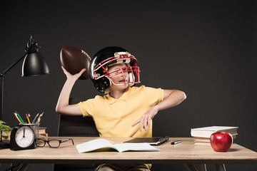 schoolboy in american football helmet throwing ball and sitting at table with books, plant, lamp,...