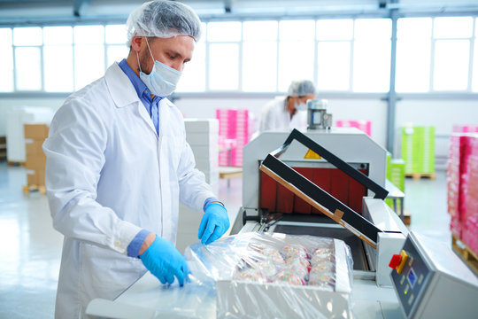 Side View Of Confectionery Factory Worker In White Coat Using Machinery To Pack Paper Box With Pastry Into Plastic Film.