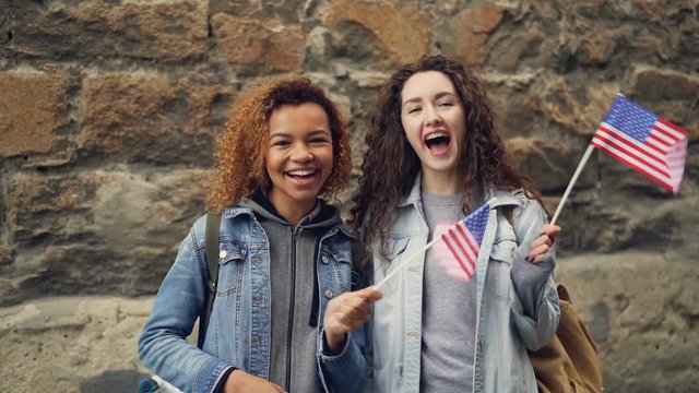 Slow motion portrait of two friends young women in casual clothes waving American flags and laughing looking at camera. Friendship, tourism and happy people concept.