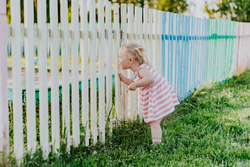 A little funny girl looks through the fence to the playground