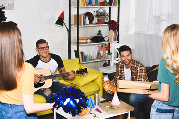 smiling young men playing guitar and looking at girls holding pizza at home party