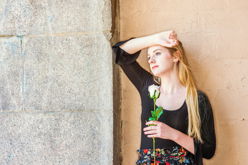 Young American teenage girl with long blonde hair, white skin, missing you, thinking about you, holding white rose, standing by wall on street in New York, arm resting on forehead, looking away..