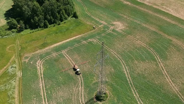 Agriculture tractor spraying rapeseed field with chemicals near high electricity tower, aerial view