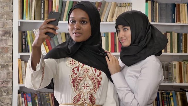 Young black muslim women in hijab with white friend taking selfie in library, smiling and happy, islamic students