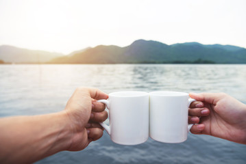 Relaxed Couple hands holding cups of hot coffee with river and mountain view.