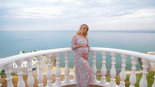 A successful pregnant girl is standing on the balcony against the sea in the evening and stroking her belly. The beginning of a new life.