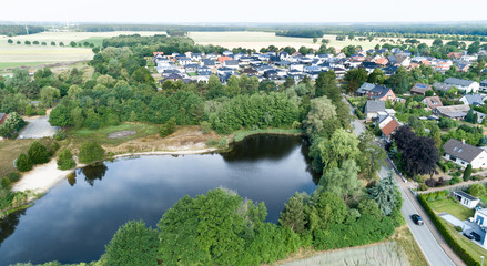 Fototapeta na wymiar Aerial view of a suburb behind a small lake and a wooded area, with detached houses, semi-detached houses and terraced houses with small front gardens and green lawns in northern Germany