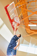 From below shot of young sportsman in basketball uniform trying to shoot goal while playing in gym. 