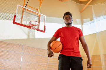 Handsome African-American man holding ball and looking at camera while standing on basketball court in gym. 
