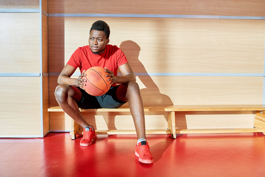 African-American sportsman holding basketball ball and looking away while resting on bench in gym. 