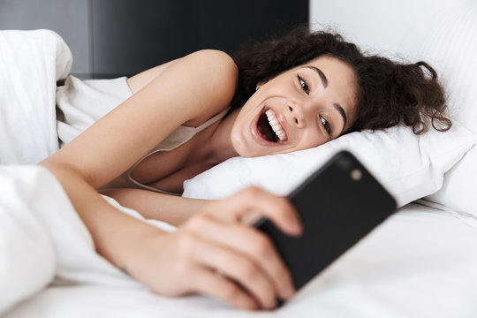 Image of joyful caucasian woman 20s with dark curly hair lying in white bed at home, and looking at mobile phone with excitement