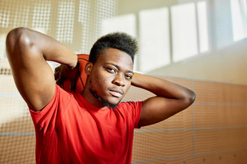 Black sportsman holding basketball ball behind head and looking at camera while standing in gym. 