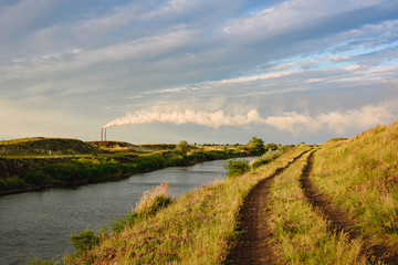 Landscape ecology and production of power, evening view of the green field and water canal, in the background smoke from the pipes of a coal-fired power station.