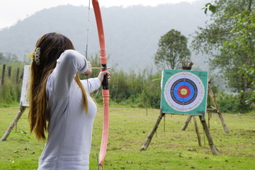 Young woman is aiming in archery  practice n the field with a target in front of her. - obrazy, fototapety, plakaty