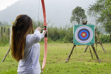 Young woman is aiming in archery  practice n the field with a target in front of her.