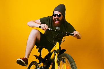 Fototapeta na wymiar Cheerful bearded hipster man ride a bike over yellow background in studio. Smiling man with beard and wearing sunglasses