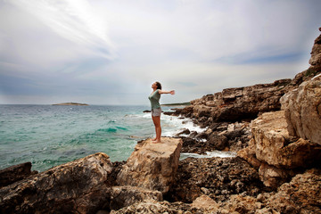 Woman with outstretched arms enjoying the wind and breathing fresh air on the rocky beach  - obrazy, fototapety, plakaty