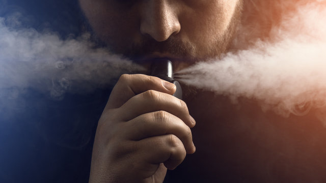 Man vape e-cigarette with e-liquid, close-up, breathes out large cloud of steam or vapor. Vaping concept, light effect