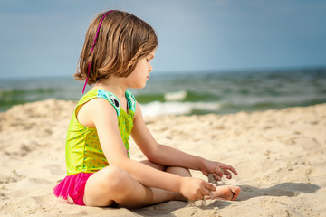 little girl in green swimsuit sitting on the sand at the beach on a summer day