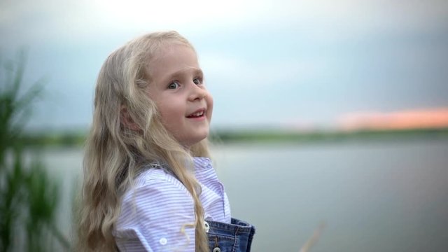 Portrait slow motion closeup of excited fisher girl 7-8 with blonde long hair fishing with wooden rod on lake or river near reeds during sunset