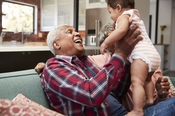 Grandparents Sitting On Sofa Playing With Baby Granddaughter At Home