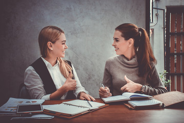 Two young european business women are sitting on the table and giving the thumbs up to agree on a decision about their work or project - obrazy, fototapety, plakaty