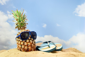 pineapple fruit with sunglasses and flip flop sandals on the beach sand against a blue sky with...