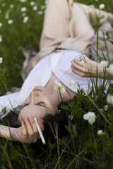 Beautiful stylish young girl wearing a trendy white shirt, beige bloomers, and a pareo lies relaxing on the green grass among the wild flowers and smoking cigarette