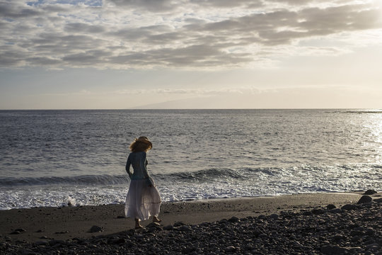 travel alone beautiful middle age lady walking on the shore at the beach. black stones and sand for vulcanic natural place. islands on the background and golden sunset light