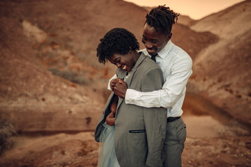 Groom hugs his bride dressed in his jacket in canyon at sunset and they laugh cheerful.