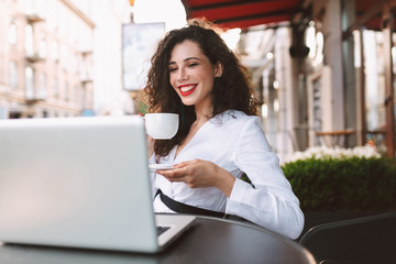 Pretty smiling woman with dark curly hair in white costume sitting at the table with cup of coffee in hands and happily looking in laptop while spending time in cafe on street