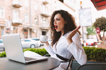 Young emotional lady with dark curly hair sitting at the table with cup of coffee in hand and amazedly looking on laptop in cafe on street