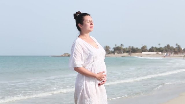 Young beautiful pregnant woman in white dress touching her belly, breathing and relax with love and care at calm blue sea on white sand. New life