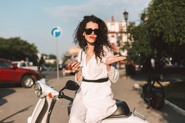 Young lady with dark curly hair in white costume and sunglasses sitting on white moped with cellphone in hand on street with city view on background