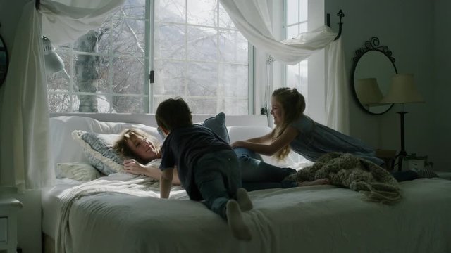 Slow motion of children waking napping mother on bed near bay window / Pleasant Grove, Utah, United States