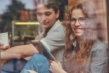 Couple of young students drinking coffee and using a digital tablet while sitting on a window sill at a college campus during a break.