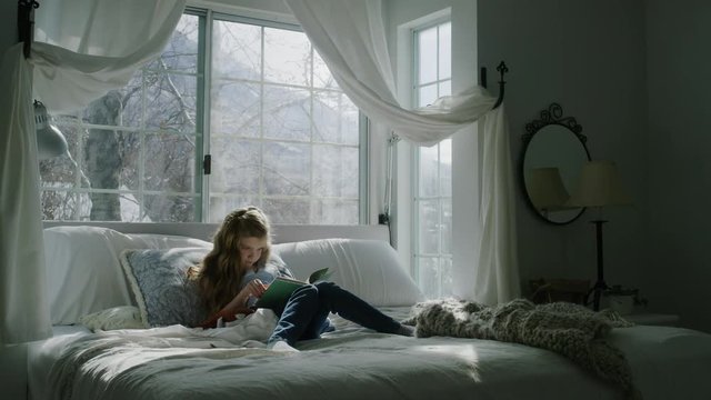 Tilt up of girl sitting in bed reading book near bay window / Pleasant Grove, Utah, United States