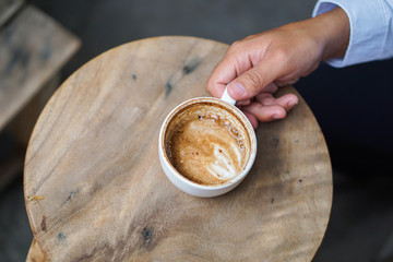 Man hand holding Latte Art coffee cup on wooden table, He is about to raise a glass of Latte Art coffee that is placed on a circular wooden table to drink.