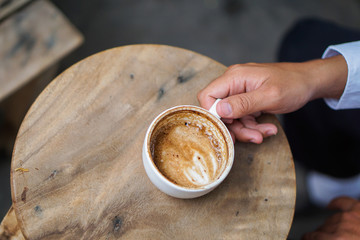 Man hand holding Latte Art coffee cup on wooden table, He is about to raise a glass of Latte Art coffee that is placed on a circular wooden table to drink.