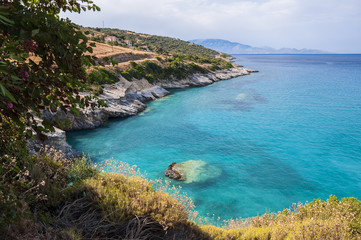 Fototapeta na wymiar A beautiful summer seascape of bay with crystal turquoise water and rocky coastline with some green flora. Colorful, vacation landscape of greek sea.