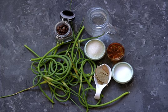 Ingredients for marinating fresh young garlic arrows on a dark gray concrete background. Fresh garlic hands, salt, sugar, vinegar, bay leaf, water, sweet pepper.