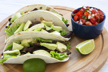 Side view, corn tortillas with grilled beef, avocado, lime and salsa on wooden board, close-up.