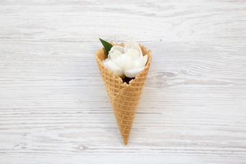 Sweet waffle cone with white peony flowers over white wooden background, top view.