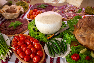 A large piece of white natural cheese on a wooden table near pickled tomatoes and cucumbers. Meat cut on a wooden stand.