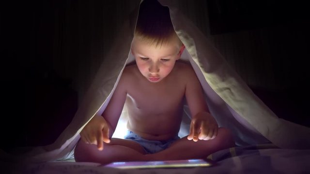 Young blond boy under covers playing on tablet a computer game.