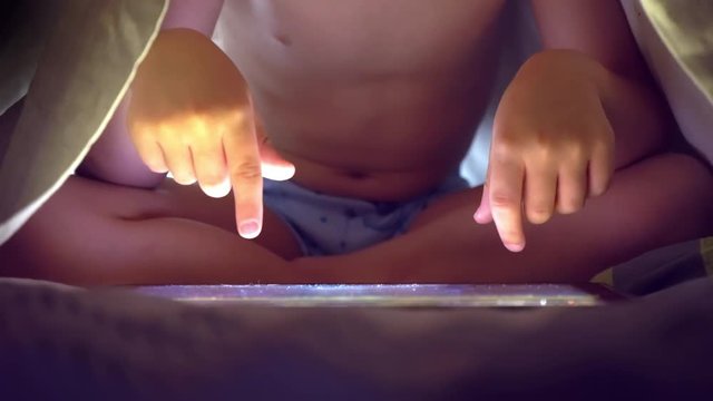 Young blond boy under covers playing on tablet a computer game.