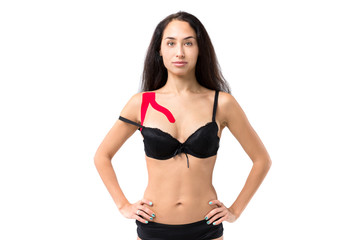 Fototapeta premium The theme kinesiology taping, help stretching, trauma and muscle pain. Young caucasian woman with long brunette hair posing in black lingerie with red lipstick tape on shoulder on isolated background