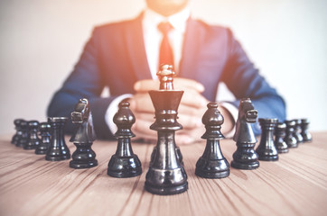 Retro style image of a businessman with clasped hands planning strategy with chess figures on an old wooden table. - obrazy, fototapety, plakaty