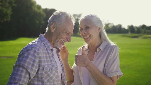 Portrait of funny elderly couple laughing at a joke outdoors. Seniors during vacation enjoying and laughing together on green lawn. Attractive woman with gray hair and handsome bearded male enjoying