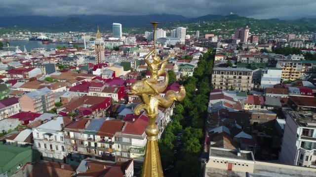 Aerial camera flying around gold figure boy riding a dolphin with a horn on the tip of the spire of the tower with an astronomical clock in Batumi on the square of Europe. Beautiful panoramic view.